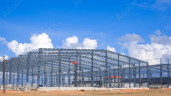 Fototapeta Metal structure of large factory warehouse industry buildings with construction workers are working in construction site against clouds on blue sky background