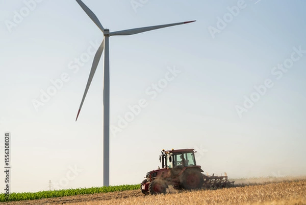 Fototapeta Agricultural tractor is working on the field with wind turbines