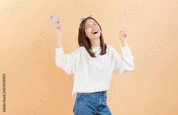 Fototapeta Cheerful beautiful Asian woman holding credit card isolated on beige color background.