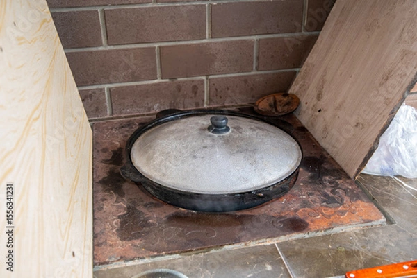 Fototapeta Large cauldron with a steaming lid simmers in a brick oven built into the wall with wood paneling on the sides, creating a cozy and rustic cooking space.