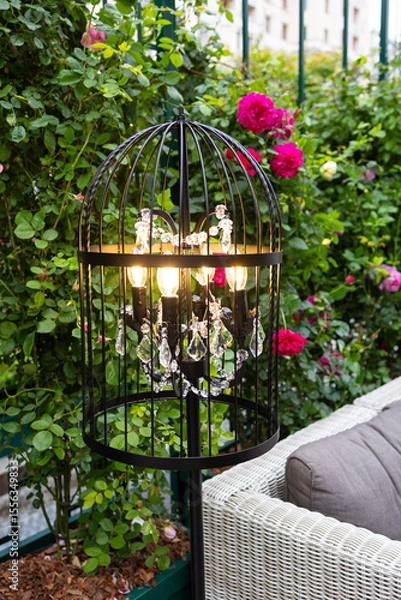 Fototapeta Decorative lantern in the garden. The lantern is made in a black checkered color and is surrounded by bright pink roses and green foliage.