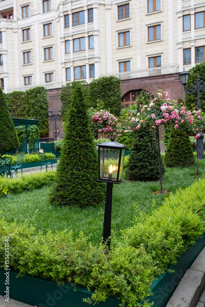Fototapeta Decorative lantern in the garden. The lantern is made in a black checkered color and is surrounded by bright pink roses and green foliage.