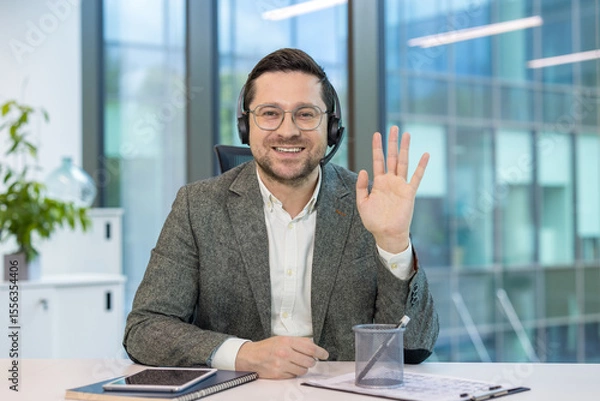 Fototapeta Portrait of a businessman in a headset and suit sitting at a desk in the office, smiling and waving at the camera