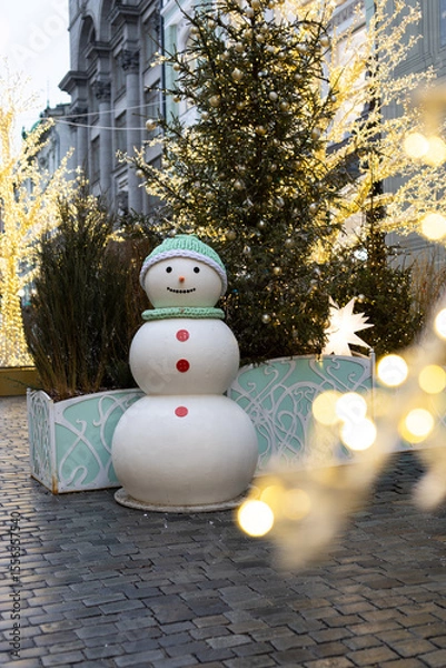 Fototapeta A cheerful snowman stands in a festive urban setting. Decorated with elegant Christmas trees and twinkling lights, creating the atmosphere of a winter holiday.
