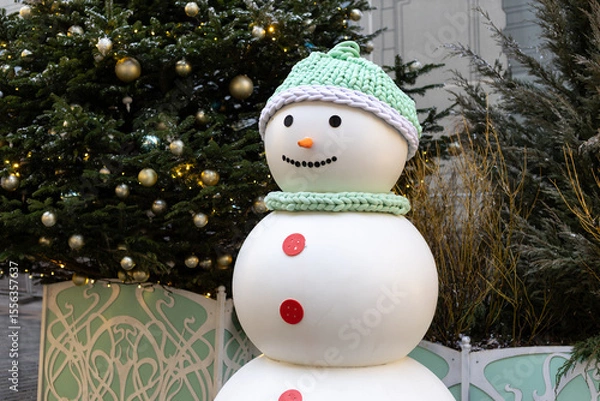 Fototapeta A cheerful snowman stands in a festive urban setting. Decorated with elegant Christmas trees and twinkling lights, creating the atmosphere of a winter holiday.
