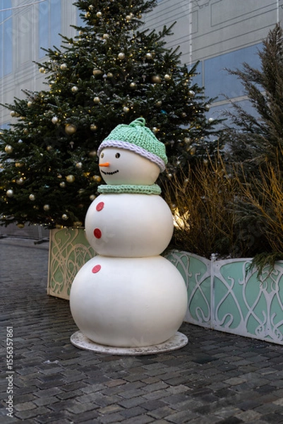 Fototapeta A cheerful snowman stands in a festive urban setting. Decorated with elegant Christmas trees and twinkling lights, creating the atmosphere of a winter holiday.