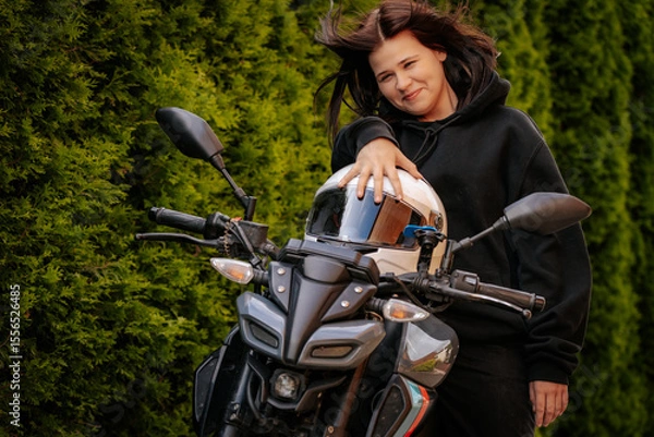 Fototapeta Young woman in black hoodie smiles while sitting on a motorcycle, set against a backdrop of lush green hedges on a sunny day.