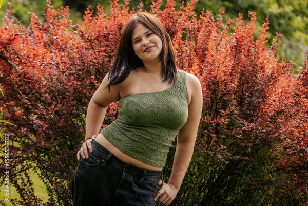 Fototapeta Smiling young woman in a textured green one-shoulder top poses in front of vibrant red foliage, wearing a necklace with an initial pendant.