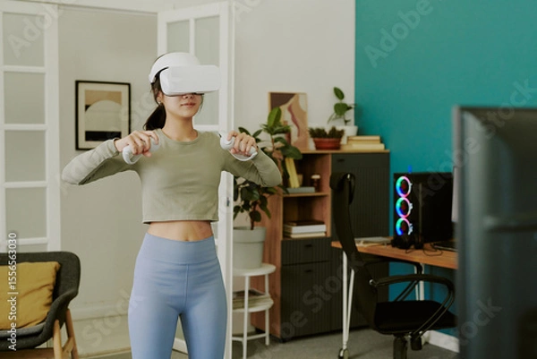 Obraz Teenage girl standing in modern room using VR headset and controllers, engaging in virtual reality exercise or gaming, computer setup with colorful lights visible in background