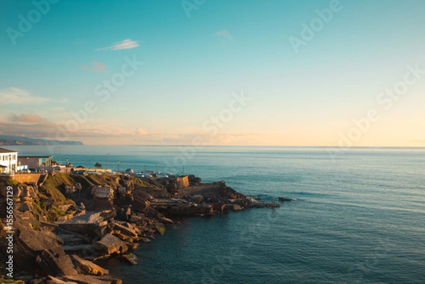 Obraz Golden hour sunlight bathes the rocky coastline of Ericeira, Portugal, as the calm Atlantic Ocean stretches out to the horizon under a tranquil sky