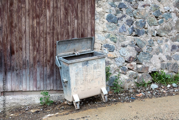 Fototapeta Old dirty metal trash bin with open lid standing on a muddy street near stone and wooden wall, concept of pollution and urban decay