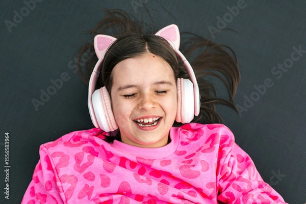 Fototapeta Smiling girl lying down with eyes closed, wearing pink headphones with cat ears and pink leopard sweatshirt, enjoying music, concept of childhood and joy