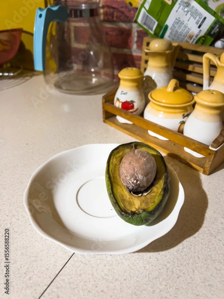 Obraz Sliced, rotten avocado with a pit on a white plate, lying on the kitchen counter, in the background you can see spices and a glass jug, in natural light
