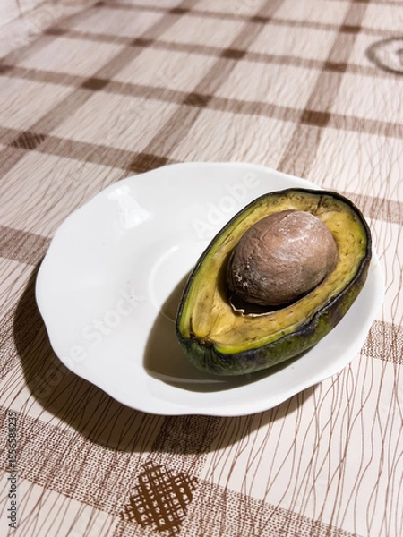 Obraz Rotten avocado with a large pit, lying on a white plate on a patterned tablecloth, showing the waste of food and staleness