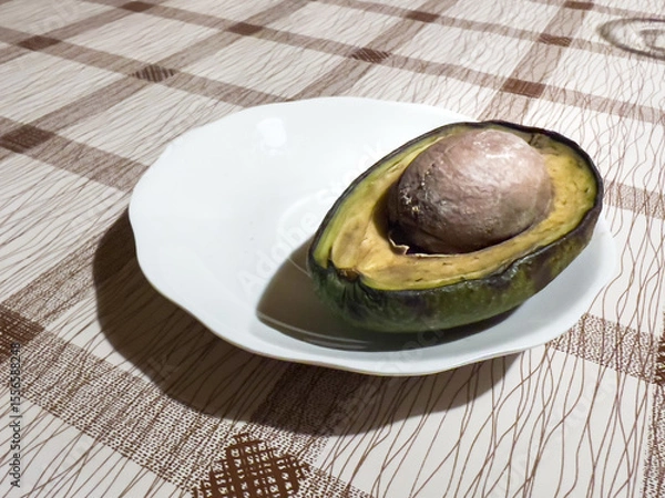 Obraz A single half of a rotten avocado with a large pit, placed on a white plate, resting on a brown and beige grid tablecloth