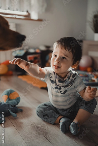 Obraz A joyful toddler sitting on a wooden floor, reaching towards the dog, in a bright, homely setting full of toys