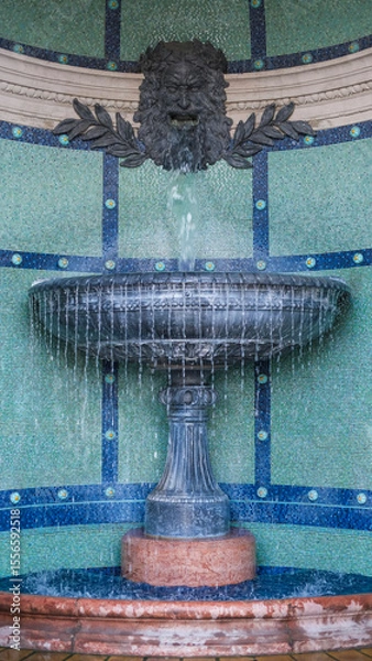 Fototapeta Decorative stone fountain with a lion-faced spout pouring water into a tiled basin, set against a mosaic wall in Budapest.