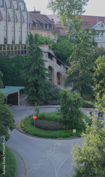 Fototapeta Stone medieval-style structure with arched entrances nestled in lush greenery near a modern building in Budapest.