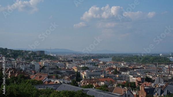 Fototapeta Panoramic view of Budapest rooftops and the Danube River stretching toward the horizon, under a lightly clouded sky.