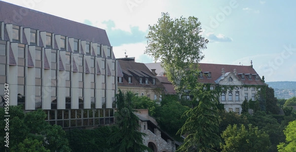 Fototapeta Contrasting architectural styles in Budapest: a modern glass building alongside historic baroque and neoclassical structures, surrounded by lush greenery.