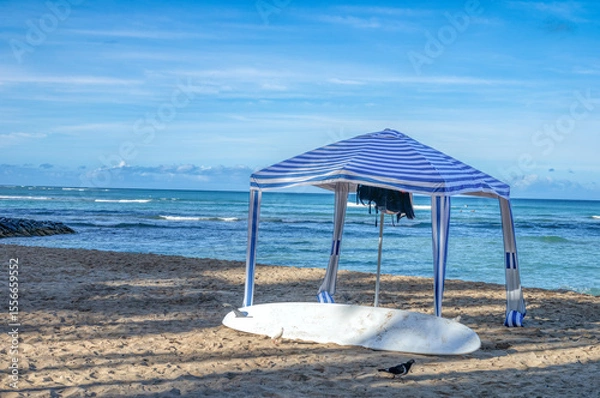 Fototapeta Blue and White Beach Cabana with a White Surfboard on an Empty Ocean and Beach.