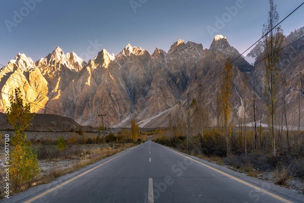 Obraz A straight, empty road stretches toward rugged, snow-dusted peaks glowing in golden hour light, flanked by tall trees and distant power lines.