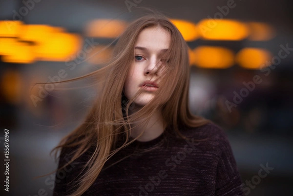 Fototapeta A moody portrait photograph of a young woman with long straight brown hair blowing in the wind against a blurred background of orange bokeh lights. 