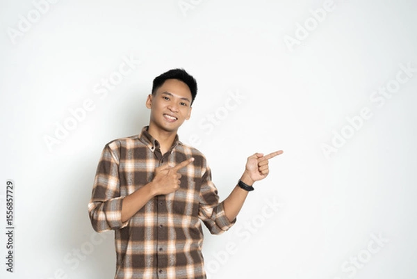 Fototapeta A cheerful man in a plaid shirt standing against a white background, pointing toward a direction. Ideal for concepts portraying direction, guidance, and cheerful expressions.
