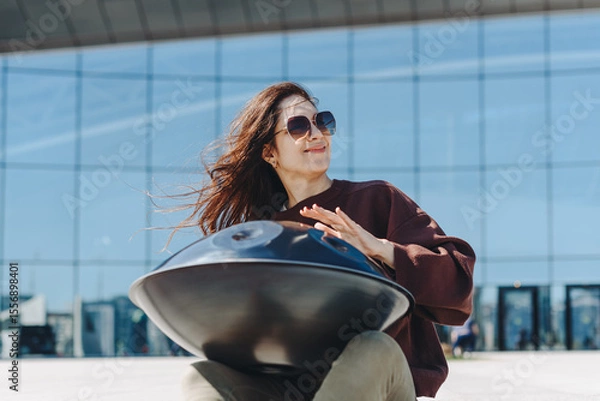 Fototapeta young caucasian woman playing black handpan with hands, ethnic metal drum instrument, sits on stone staircase, scyscraper and business center in background, beautiful relaxing music for meditation