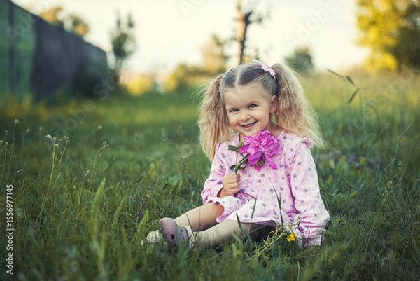 Obraz cheerful 3-year-old girl with pigtails in a pink dress with a peony in a village meadow