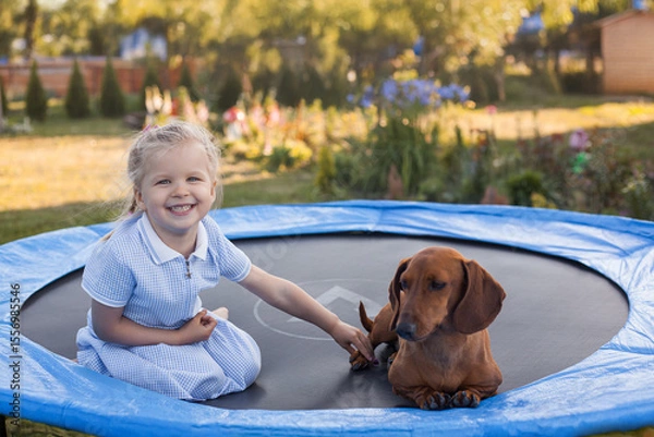 Obraz a three year old smiling girl playing with dachshund dog on trampoline in green sunny summer park