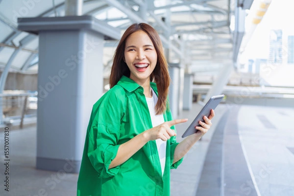 Fototapeta Smiling asian woman holding digital tablet while standing in front of modern business building