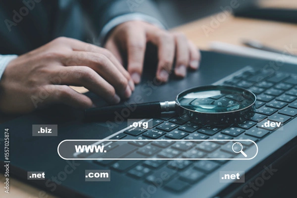 Fototapeta A close-up of hands typing on a laptop keyboard, showcasing various domain extensions such as .com, .net, and .org. The magnifying glass indicates searching for information online.
