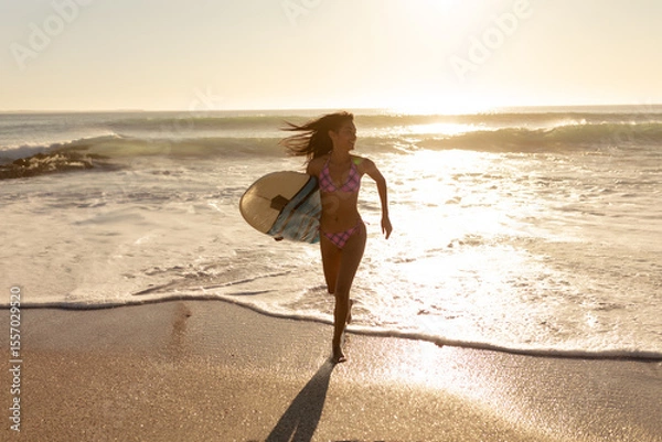 Obraz Woman running from breaking waves onto wet sand at beach edge, holding surfboard during golden hour