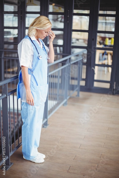 Obraz Nurse in scrubs leaning on railing holding forehead with stethoscope in mezzanine near glass doors