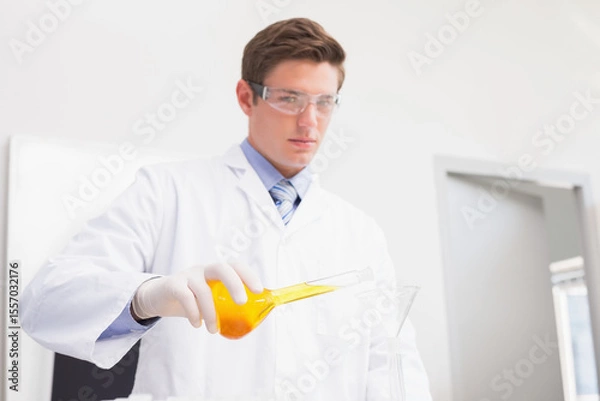 Obraz Male scientist in white coat pouring orange liquid through funnel into glass container on lab bench