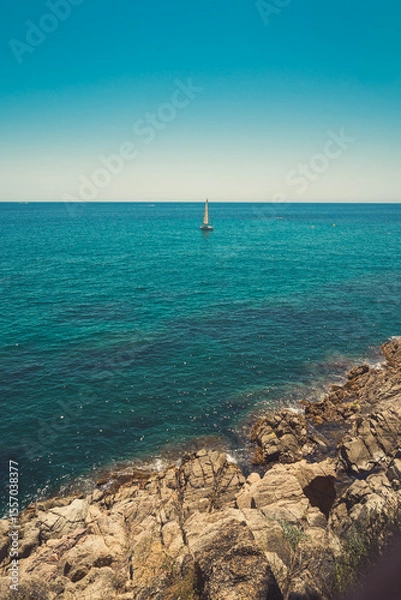 Fototapeta Seascape in Spain with a sailboat in the background and rocky cliffs in the foreground. Costa Brava coast. Vertical view.