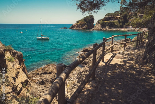 Fototapeta Seascape in Blanes, Spain with a sailboat in the background and a promenade on a cliff called Cami de Ronda in the foreground. Costa Brava coast.