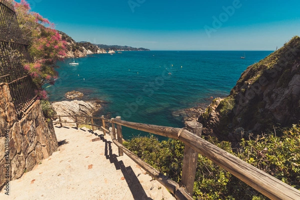 Fototapeta Seascape in Blanes, Spain with a sailboat in the background and the cliff promenade called Cami de Ronda in the foreground. Costa Brava.