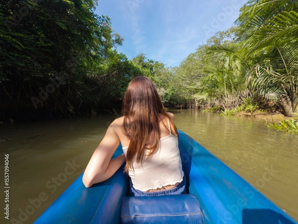 Fototapeta Rear view of a woman in a blue canoe navigating a waterway through lush Thai jungle with dense vegetation and tangled tree roots.