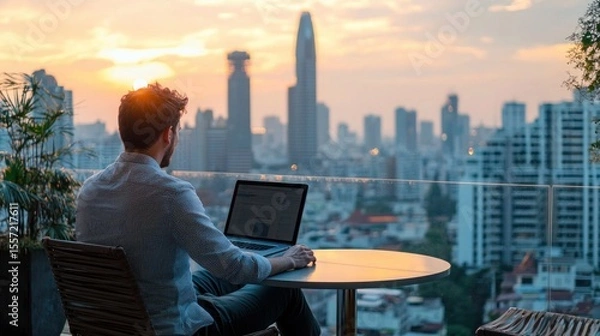 Fototapeta A travel blogger sitting on a hotel rooftop deck working on a laptop while enjoying the city skyline view