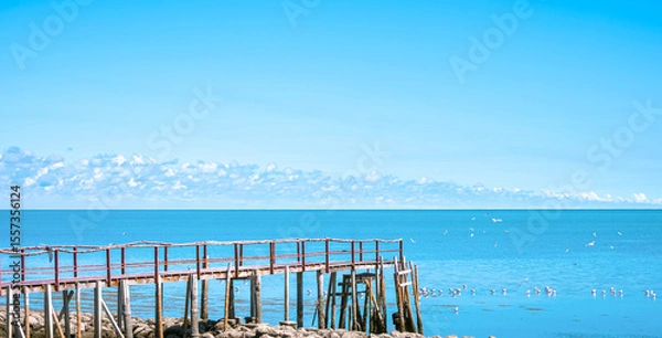 Fototapeta Wooden bridge extends into the sea in nature viewpoint area against blue summer sky background, perspective side view with copy space