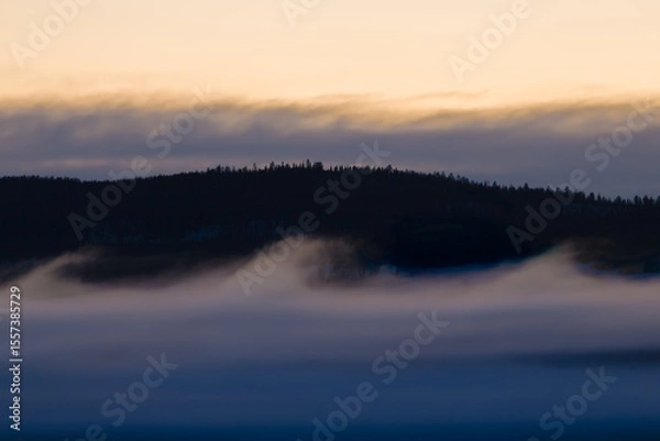 Obraz A serene mountain landscape at sunrise, with golden skies illuminating the horizon. A soft blanket of fog drifts over the dark forest below, while the snow-dusted peaks rise in the distance. 