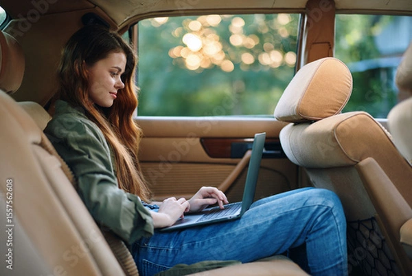 Fototapeta Young woman working on a laptop inside a car during sunset, showcasing a blend of productivity and relaxation with a serene background.