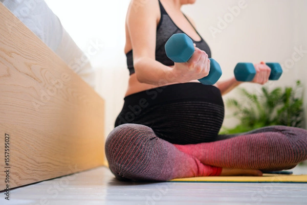 Fototapeta Serene pregnant female in cross-legged stretch pose on bed, maintaining fitness routine. Nurturing body and mind while awaiting baby's arrival.