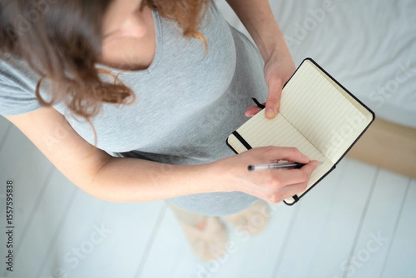 Fototapeta Overhead close-up of expecting mother carefully preparing birth plan in notebook. Organized maternity preparation and important reminders for upcoming delivery.