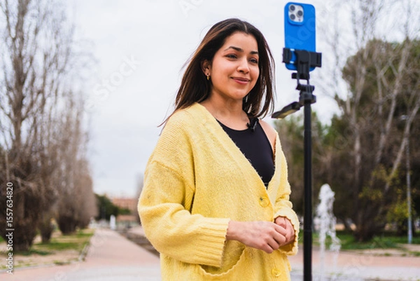Fototapeta Young woman creating online content using smartphone on tripod with lavalier microphone in a park