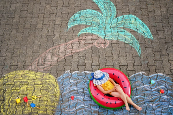 Fototapeta Happy little preschool girl in swimsuit on inflatable ring with sea, sand, palm painted with colorful chalks on asphalt. Cute child with having fun with chalk picture. Summer, vacations, summertime
