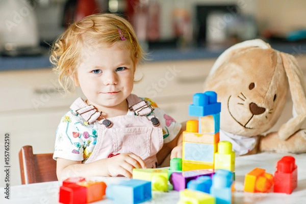 Fototapeta Adorable toddler girl with favorite plush bunny playing with educational toys in nursery. Happy healthy child having fun with colorful different plastic blocks at home. Cute baby learning creating.