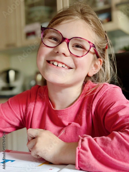 Fototapeta Cute elementary school girl sitting at a desk at home, writing in her workbook and doing school homework. Focused child learning and studying indoors. Concept of education, childhood, home learning.